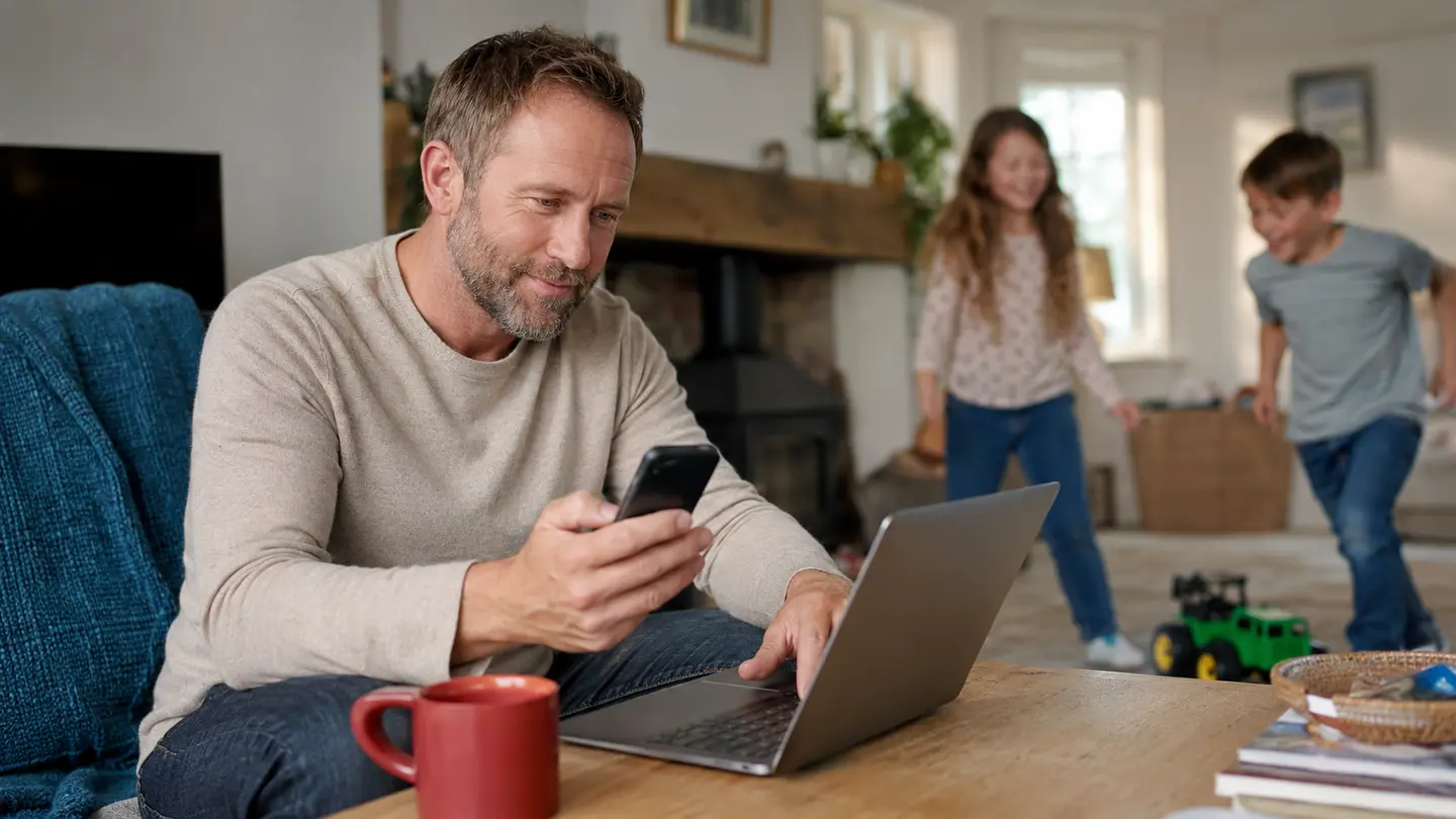 A parent backing up information on a phone and laptop while children play at home